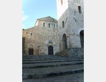 Facciata di una chiesa romanica in pietra chiara, con portali ad arco e piccole finestre, affacciata su un’ampia scalinata in pietra. A destra si vede una torre massiccia con grandi arcate e aperture strette sotto un cielo sereno.