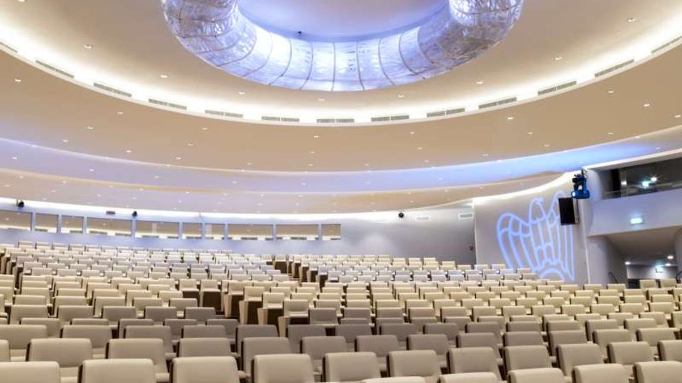 Ampia sala auditorium moderna con molte file di poltrone beige disposte a gradoni. Soffitto curvo con grande elemento circolare illuminato al centro e luci incassate lungo le pareti.