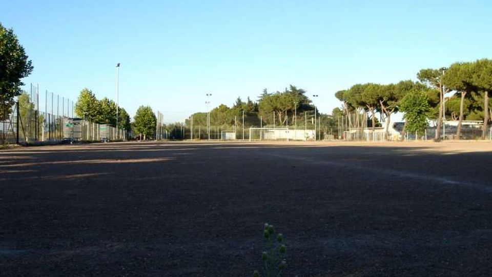 Ampio campo sportivo all’aperto con fondo scuro in terra battuta, circondato da recinzioni metalliche e pali per l’illuminazione. Sullo sfondo si vedono alberi e una piccola struttura vicino ai campi, sotto un cielo sereno.