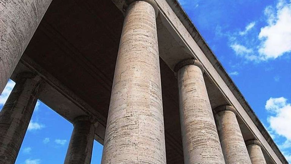 Ripresa dal basso di un viadotto in cemento armato con alte colonne cilindriche che sostengono l’impalcato superiore. Sullo sfondo si vede un cielo azzurro con nuvole sparse.