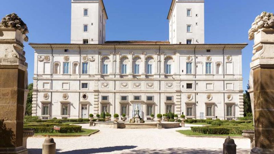 Facciata di un grande palazzo storico in stile rinascimentale, con due torri quadrate sul retro, finestre ad arco e decorazioni in stucco su più livelli. Davanti si apre un giardino formale con siepi geometriche, vialetti in ghiaia e una fontana centrale.