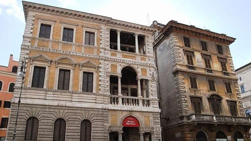 Facciata di un palazzo storico in pietra e intonaco con finestre regolari e loggia centrale ad archi e colonne, con ingresso indicato da un’insegna rossa “Museo Barracco”. Accanto si vede un edificio più stretto e usurato, su una strada urbana con lampione e cielo parzialmente nuvoloso.