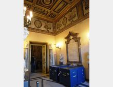 Interno di una sala storica con soffitto a cassettoni decorato e fregi dipinti lungo il perimetro, illuminata da lampadari e applique. Sulle pareti si vedono busti e monumenti marmorei, una grande lapide commemorativa e un portale con ante decorate che conduce a un ambiente adiacente.
