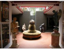 Interno di un museo o spazio espositivo con pavimento in mattoni rossi, una grande struttura circolare centrale e statue esposte su piedistalli ai lati. Sul fondo si vede una scalinata che sale verso un’apertura superiore con ringhiera e luce naturale.