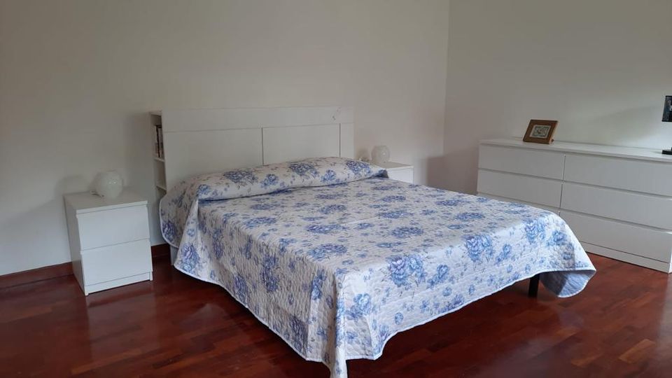 Camera da letto luminosa con pareti bianche e pavimento in legno scuro, con letto matrimoniale al centro coperto da copriletto bianco a fiori blu. Ai lati ci sono comodini bianchi e sulla parete a destra una cassettiera bianca lunga con una cornice appoggiata sopra.