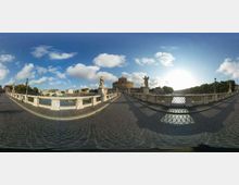 Veduta panoramica di un ponte in pietra con pavimentazione in sampietrini, balaustre e statue lungo i lati, che attraversa un fiume. Sullo sfondo si vede un grande edificio circolare fortificato e, a destra, una fila di alberi lungo la riva sotto un cielo con nuvole.