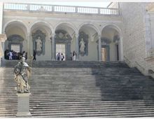 Ampia scalinata in pietra che conduce a un porticato con arcate e colonne, con nicchie decorate e statue lungo la parete. In primo piano si vede una statua su piedistallo, mentre ai lati compaiono muri in pietra dell’edificio.