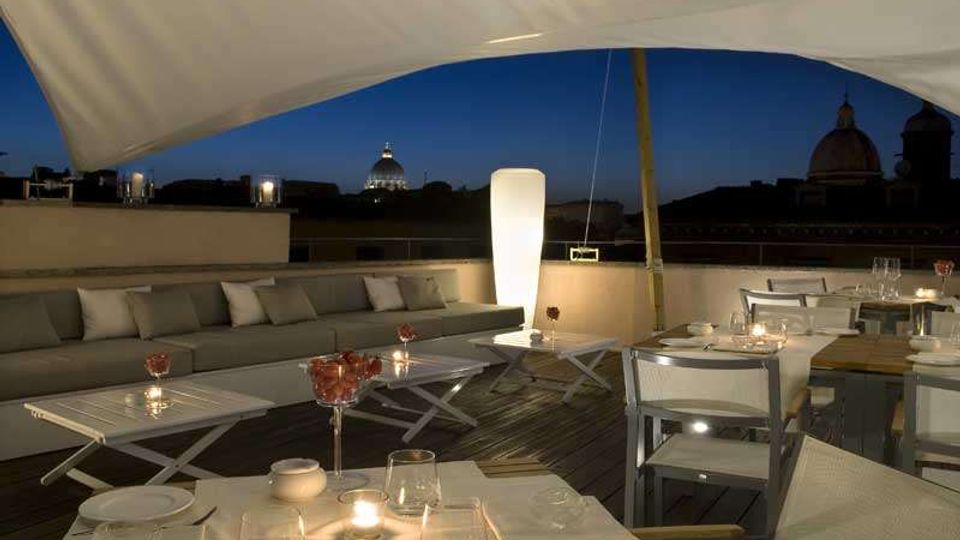 Terrazza panoramica su un tetto con tavoli apparecchiati e divani, illuminata da lampade e candele sotto una grande copertura bianca. Sullo sfondo si vede lo skyline urbano notturno con cupole e edifici storici.
