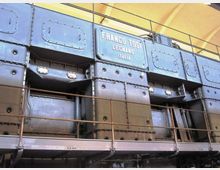 Interno industriale con grandi pannelli metallici rivettati e passerella con ringhiera in metallo, illuminato da luci incassate. Al centro si vede una targa con la scritta “FRANCO TOSI, LEGNANO, ITALIA” sotto una copertura ad arco.