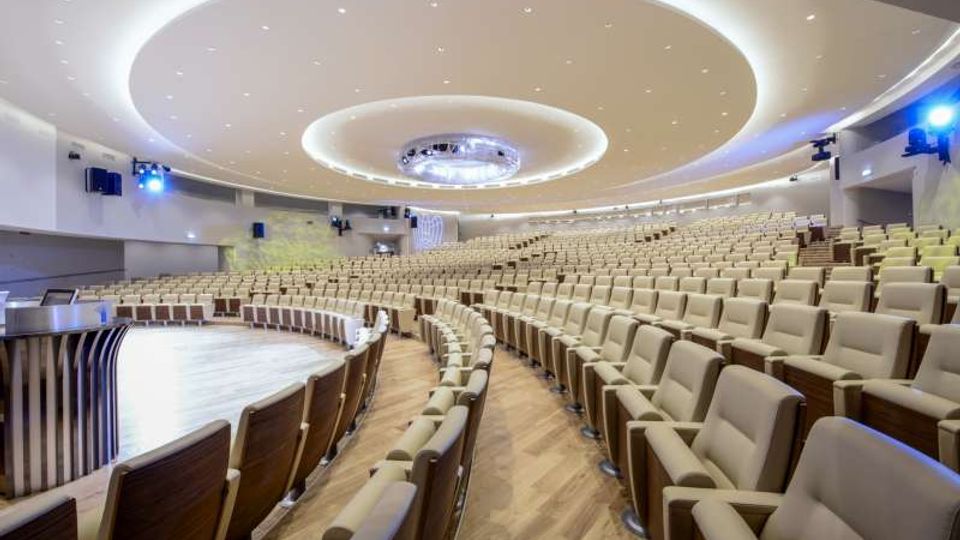 Ampio auditorium moderno con numerose file di poltrone beige disposte a semicerchio attorno a un palco curvo in legno. Il soffitto presenta grandi elementi circolari con illuminazione integrata e faretti, con pareti laterali attrezzate per audio e luci.
