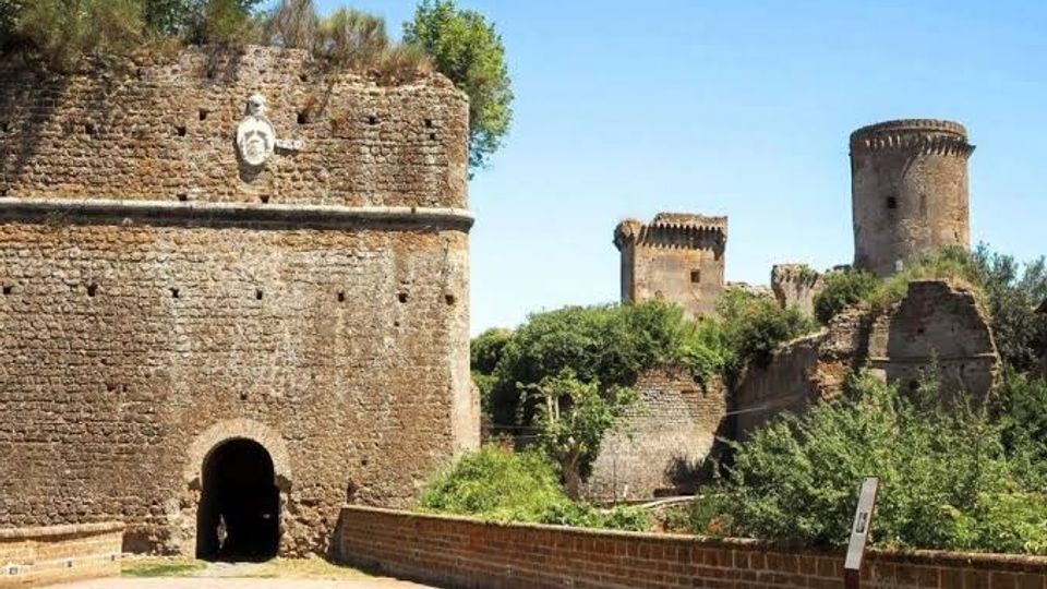 Rovine di una fortificazione in pietra con un grande muro e un arco d’ingresso, affiancata da una passerella e vegetazione fitta. Sullo sfondo si vedono torri e bastioni in pietra sotto un cielo sereno.