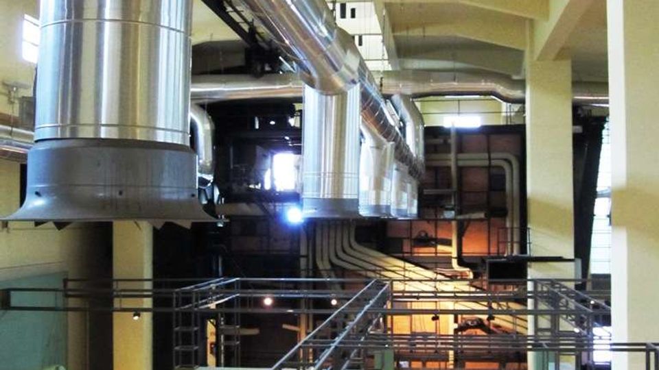 Interno di un grande edificio industriale riconvertito, con soffitto alto a travi e grandi condotti metallici di ventilazione sospesi. Passerelle e ringhiere metalliche attraversano lo spazio sopra un’area espositiva con pareti verdi e sculture su piedistalli.