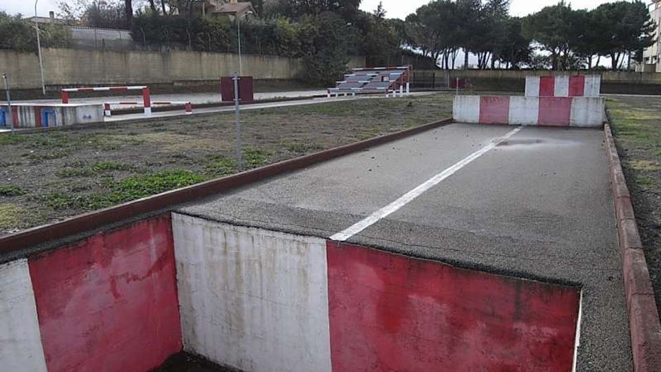 Pista asfaltata all’aperto con cordoli e barriere basse dipinte di rosso e bianco, disposta in corsie e curve come un circuito di guida. Sullo sfondo si vedono un muro di recinzione, lampioni e alberi alti lungo il perimetro.
