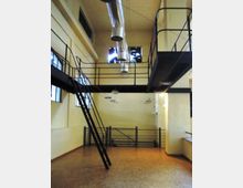 Interno di un edificio industriale riconvertito con soffitto alto, soppalco in metallo con parapetti e scala inclinata. Grandi finestre, pavimento in legno e condotte di ventilazione a vista sul soffitto.