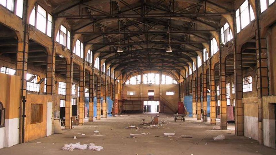 Interno di un grande capannone industriale dismesso, con struttura metallica a capriate sul soffitto e alte finestre laterali che illuminano lo spazio. Pavimento in cemento quasi vuoto con detriti sparsi e pareti in muratura con pilastri e impalcati laterali.