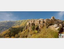 Borgo medievale in pietra arroccato su una rupe, con case addossate l’una all’altra e una chiesa con campanile. Intorno si estende una valle boscosa con colline alberate sotto un cielo azzurro.