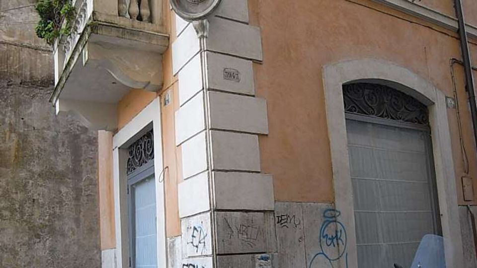 Angolo di strada in un centro storico con edificio intonacato color ocra e pietra bugnata, finestre con persiane e una piccola balconata con piante. Al piano terra si vedono porte e vetrine con grate, muri con graffiti e alcune biciclette e scooter parcheggiati lungo i paletti.