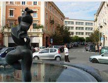 Fontana circolare in una piazza urbana, con una grande statua scura al centro e vasca d’acqua in primo piano. Sullo sfondo si vedono palazzi multipiano con facciate color pastello, strada con auto parcheggiate e alcuni alberi lungo i marciapiedi.