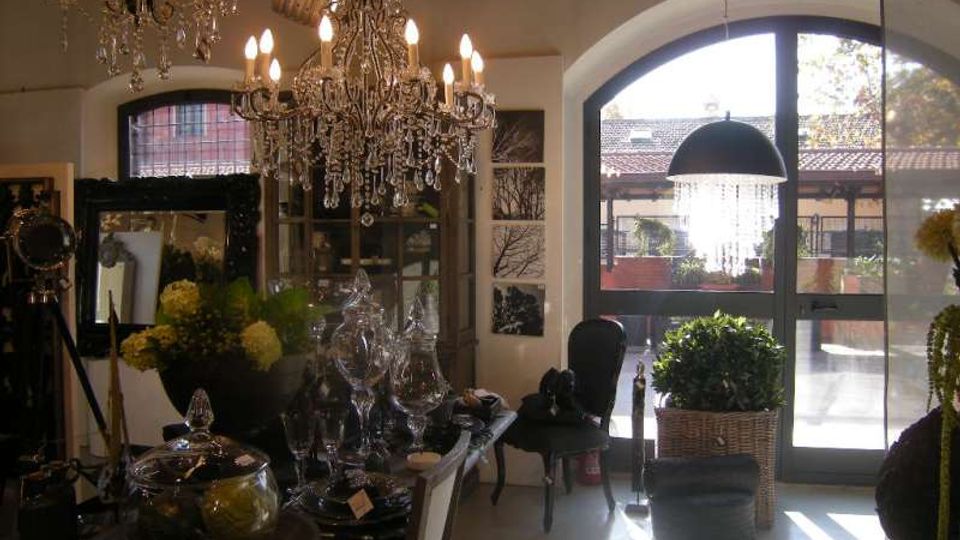 Interno di un negozio o showroom con lampadari in cristallo appesi al soffitto e tavoli espositivi con oggetti decorativi e vasi. Grandi porte-finestre ad arco illuminano l’ambiente e si affacciano su un cortile esterno con edifici e piante.