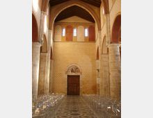 Interno di una chiesa in stile romanico, con navata centrale fiancheggiata da colonne in pietra e archi a tutto sesto. In fondo si vede una grande porta in legno con lunetta affrescata sopra, pareti in laterizio e piccole finestre alte, e pavimento a mosaico geometrico con file di sedie trasparenti ai lati.