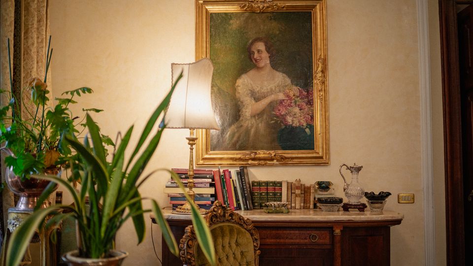 Interno di una stanza in stile classico con grande ritratto incorniciato in oro sopra una console in legno con piano in marmo. Sulla console ci sono una lampada con paralume, libri e oggetti decorativi, con piante in vaso e una sedia imbottita in primo piano.