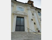 Facciata di un edificio storico color crema con cornici e lesene bianche decorate, grande portale con grata metallica e scalinata in pietra con corrimano. In alto si vede una finestra ad arco e una torre campanaria parzialmente visibile sul tetto.