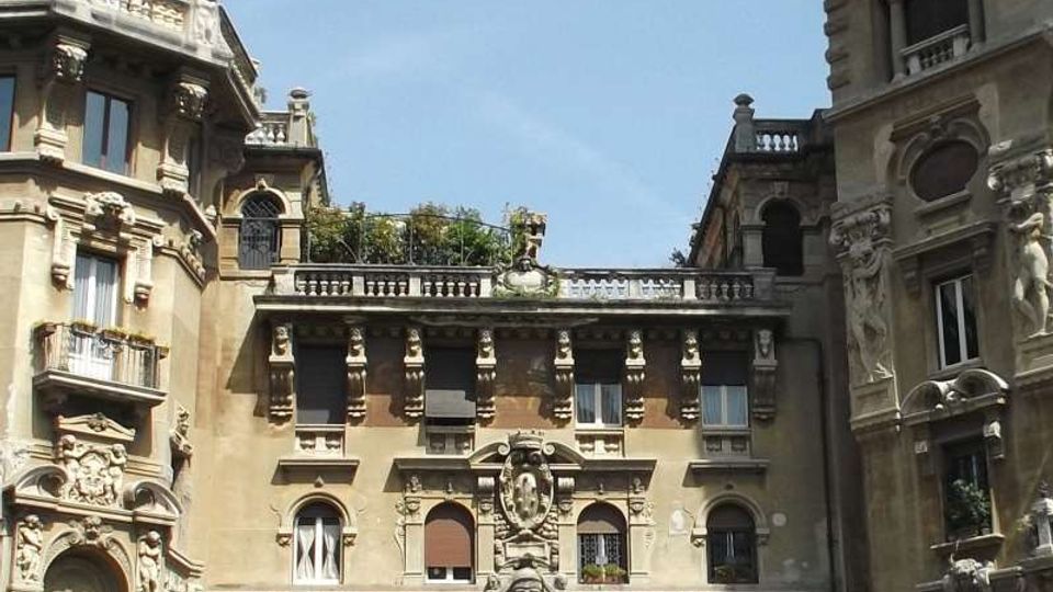 Veduta stradale urbana con palazzi storici in pietra riccamente decorati e un grande arco monumentale che collega due edifici sopra la carreggiata. Si notano balconi, finestre con cornici ornate e sculture sulla facciata, con automobili parcheggiate e cielo sereno sullo sfondo.