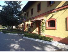Edificio a due piani con facciata giallo ocra e cornici rosse intorno a finestre e porte, con piccolo portico d’ingresso. Davanti c’è un vialetto pavimentato con aiuole bordate di bianco, bandiere su aste e alberi che proiettano ombre sul suolo.