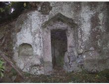 Ingresso rettangolare scavato nella roccia, con architrave a forma di timpano e pareti in pietra grezza. La facciata è coperta da licheni e muschio, inserita in un’area boscosa con vegetazione ai margini.