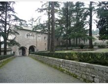 Viale in pietra con pavimentazione a ciottoli che conduce a un edificio storico in muratura con archi d’ingresso e finestre ad arco. Ai lati si vedono muretti in pietra, siepi basse e alti alberi che circondano il complesso.