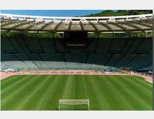 Interno di uno stadio all’aperto con ampie tribune a gradoni e copertura superiore, visto dal lato del campo. In primo piano si vede il prato verde con le linee di gioco e una porta da calcio, circondato da una pista di atletica.