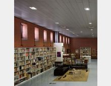 Interno di una biblioteca o sala lettura con pareti in mattoni rossi, alte finestre strette e soffitto a pannelli con illuminazione a incasso. Lungo la parete si estendono scaffali bianchi pieni di libri, con un’area lounge centrale con divani scuri, tavolini e tappeto.
