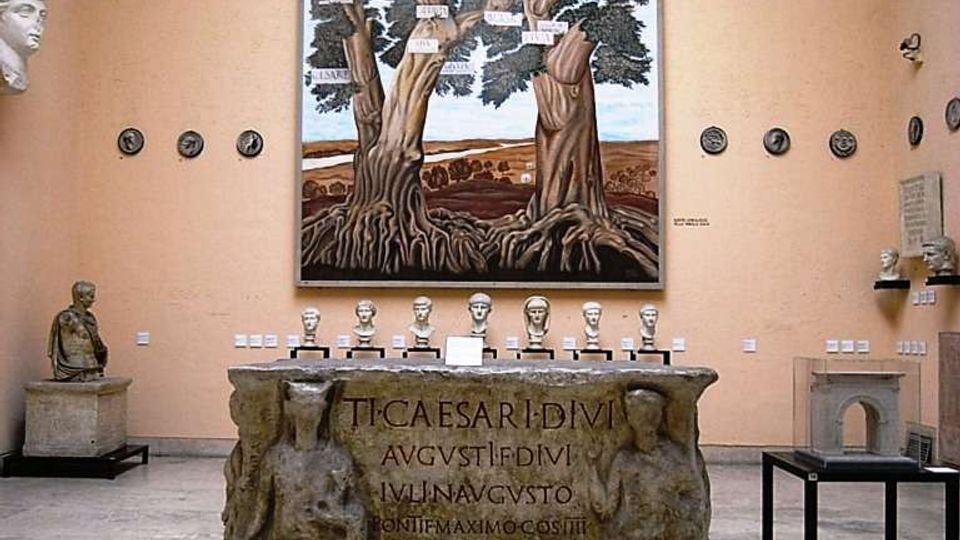 Sala museale con pareti color pesca e pavimento in pietra, con al centro un grande altare lapideo inciso e affiancato da reperti esposti su piedistalli e teche. Sulla parete di fondo è appeso un grande dipinto raffigurante un albero, con medaglioni e busti antichi disposti lungo le pareti.