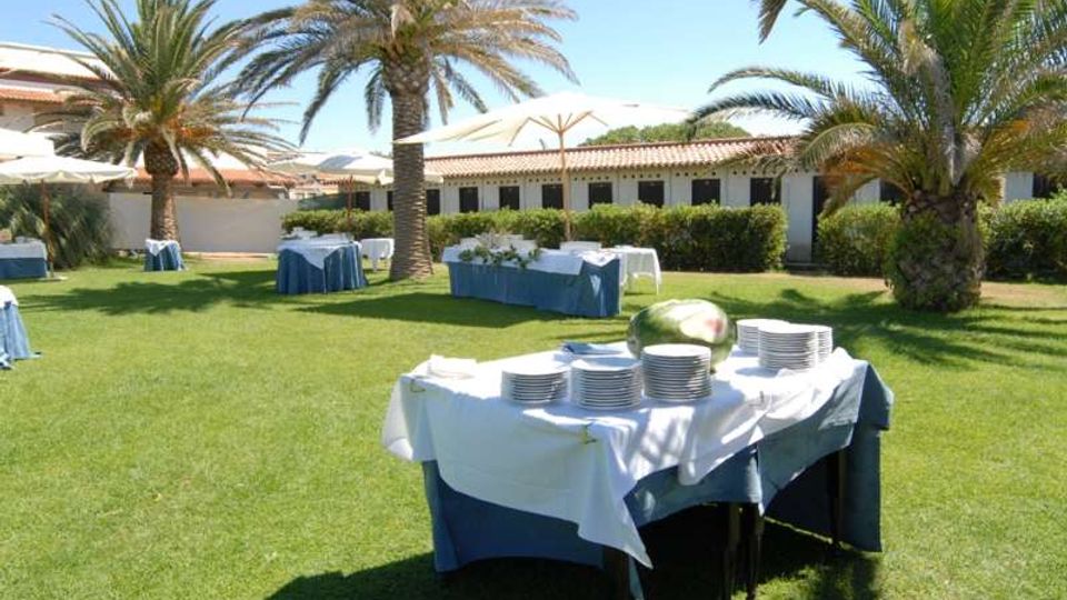 Giardino con prato verde e palme, allestito per un ricevimento con tavoli coperti da tovaglie bianche e blu, piatti impilati e ombrelloni. Sullo sfondo si vede un edificio basso con tetto in tegole e siepi lungo la facciata.