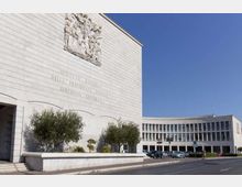Complesso di edificio istituzionale in pietra chiara con grande facciata liscia, bassorilievo e iscrizione incisa; sullo sfondo un’ala curva con file di finestre. Davanti si vedono un’aiuola con alberi, un parcheggio con auto e una strada sotto cielo sereno.