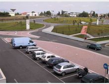 Rotatoria stradale moderna con aiuole erbose e giovani alberi al centro, circondata da carreggiate asfaltate e cartelli direzionali. In primo piano si vede un parcheggio con diverse auto e un furgone, con edifici bassi sullo sfondo.
