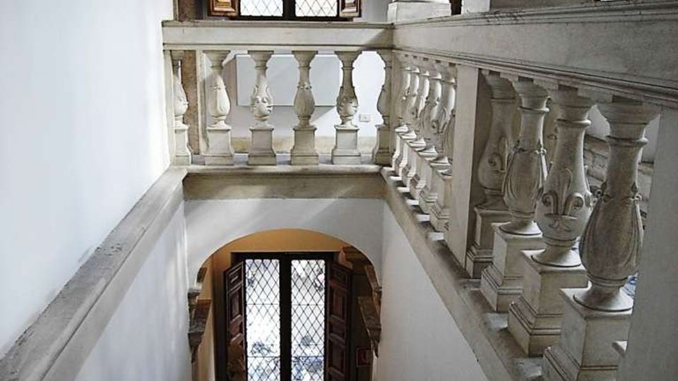 Interno di un edificio storico con scala in pietra che scende verso un pianerottolo, fiancheggiata da una balaustra in marmo e corrimano in ferro. In alto si vedono grandi finestre con griglie e persiane in legno, e colonne scure che sostengono il piano superiore.