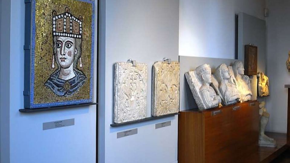 Sala museale con pareti bianche che espone un grande mosaico a tessere dorate con ritratto frontale e, accanto, rilievi in pietra appesi e busti marmorei allineati su un lungo basamento in legno. Illuminazione puntuale sulle opere e pavimento in legno.