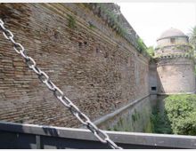 Mura di una fortificazione in mattoni viste da un ponte con parapetto e una grossa catena metallica in primo piano. Sullo sfondo si nota una torre cilindrica con cupola e vegetazione lungo il fossato.