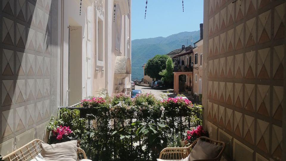 Piccola terrazza con sedute in rattan e tavolino su pavimento a motivi geometrici, delimitata da una ringhiera con fiori. L’area è incorniciata da un grande arco tra due pareti decorate e si affaccia su una strada del borgo con montagne sullo sfondo.