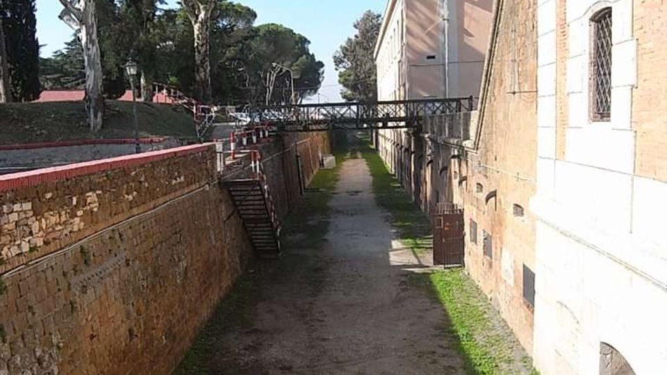 Fossato asciutto di una fortificazione, con percorso in terra e tratti d’erba tra alte mura in mattoni e pietra. In fondo si vede un ponte pedonale in ferro; sul lato sinistro ci sono alberi e una scalinata che scende nel fossato.