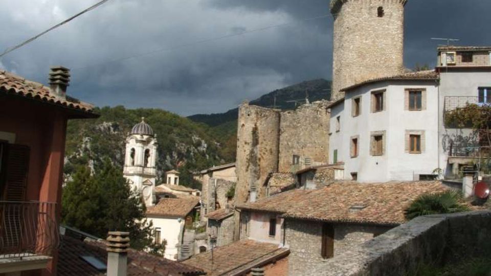 Veduta di un borgo in pietra con tetti in coppi, una torre cilindrica medievale e resti di mura, affiancati da edifici intonacati con finestre. Sullo sfondo si vedono colline boscose e un cielo nuvoloso.