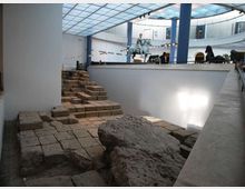 Interno di un museo con copertura in vetro e colonne, affacciato su un’area archeologica ribassata con pavimentazione in pietra, gradini e grandi blocchi rocciosi. Su una piattaforma superiore si intravedono sculture, tra cui una statua equestre, lungo il bordo del vuoto centrale.