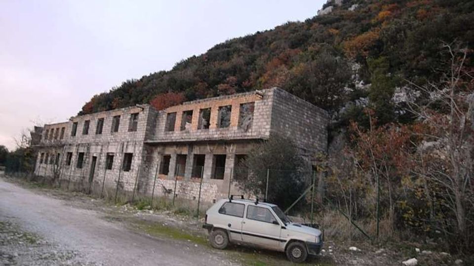 Edificio rettangolare in muratura grezza a due piani, con file di finestre vuote e portico frontale, situato lungo una strada sterrata. Sullo sfondo si vede un pendio collinare boscoso e roccioso, con vegetazione fitta e recinzione metallica vicino alla struttura.
