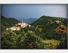 Veduta panoramica di un borgo arroccato con case in pietra e campanile, circondato da colline boscose e vallate verdi. Sullo sfondo si intravedono montagne e un cielo nuvoloso.