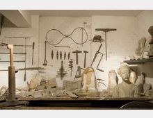 Interno di un laboratorio di scultura con parete bianca su cui sono appesi utensili e sagome metalliche. In primo piano un banco da lavoro ingombro di frammenti e busti in gesso, con scaffali laterali pieni di modelli e materiali.