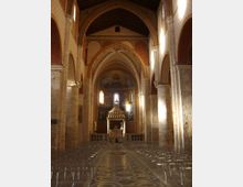 Interno di una chiesa in stile romanico-gotico con navata centrale, alte arcate in pietra e soffitto a capriate in legno. In fondo si vede l’altare sotto una struttura a baldacchino, affreschi sull’abside e file di sedie disposte lungo il pavimento decorato a motivi geometrici.