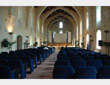 Interno di una grande sala conferenze ricavata in un edificio in pietra con alte arcate e soffitto a capriate, illuminata da finestre ad arco sui lati. File di poltrone blu sono orientate verso un palco con grande schermo di proiezione e pannelli informativi sullo sfondo.