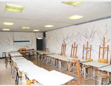 Aula di laboratorio artistico con tavoli bianchi disposti in file e sgabelli, illuminata da pannelli luminosi a soffitto. Lungo la parete destra ci sono cavalletti in legno e pareti bianche macchiate di schizzi di colore.