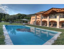Piscina all’aperto con bordo in pietra su prato, davanti a una grande villa color ocra con portico ad archi, pilastri e tetto in legno a vista. Sullo sfondo si vedono alberi e colline verdi sotto un cielo sereno.